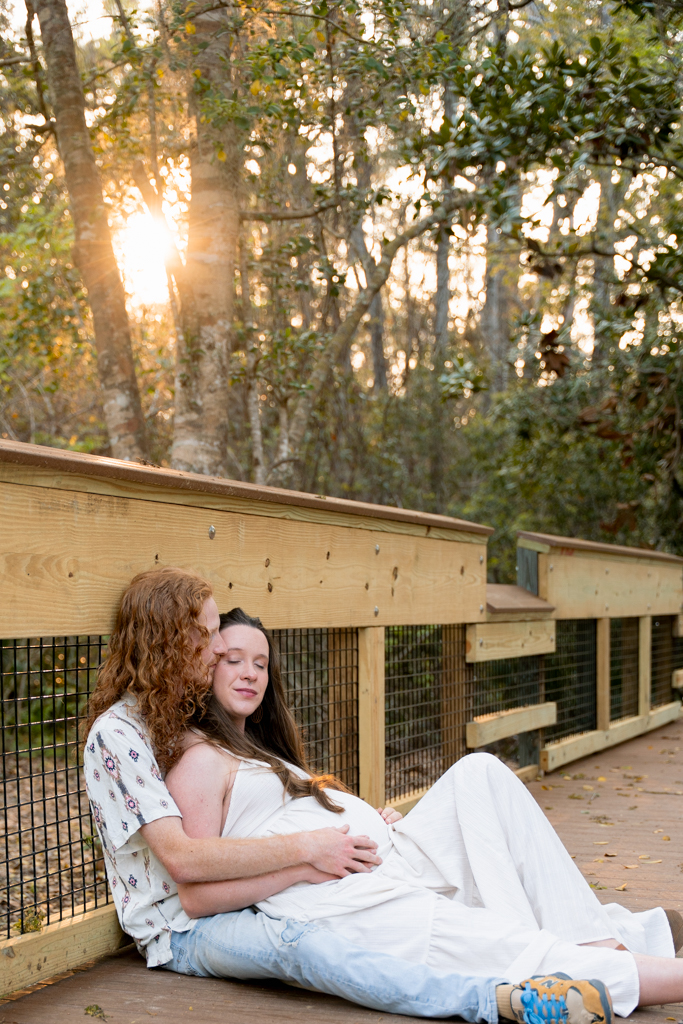 Pregnant woman twirling in a light dress under the shade of tall oak trees at Eden Gardens.