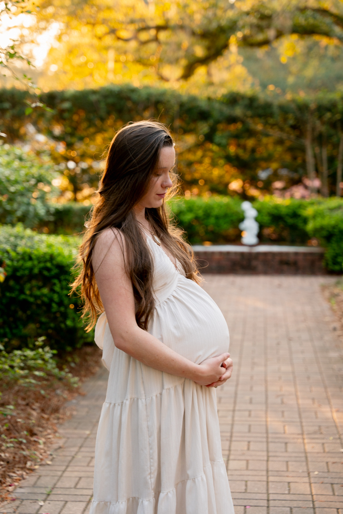 Pregnant woman in flowing white dress standing beneath moss-draped oak trees at Eden Gardens State Park.