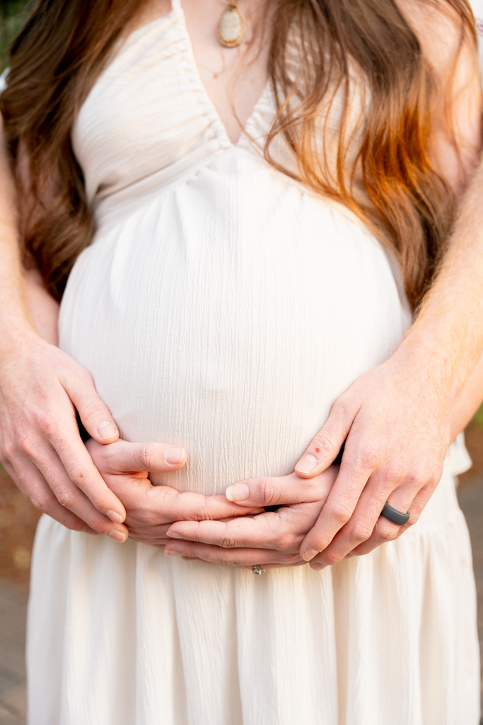 Close-up of hands forming a heart shape over a baby bump in a lush garden setting at Eden State Park.