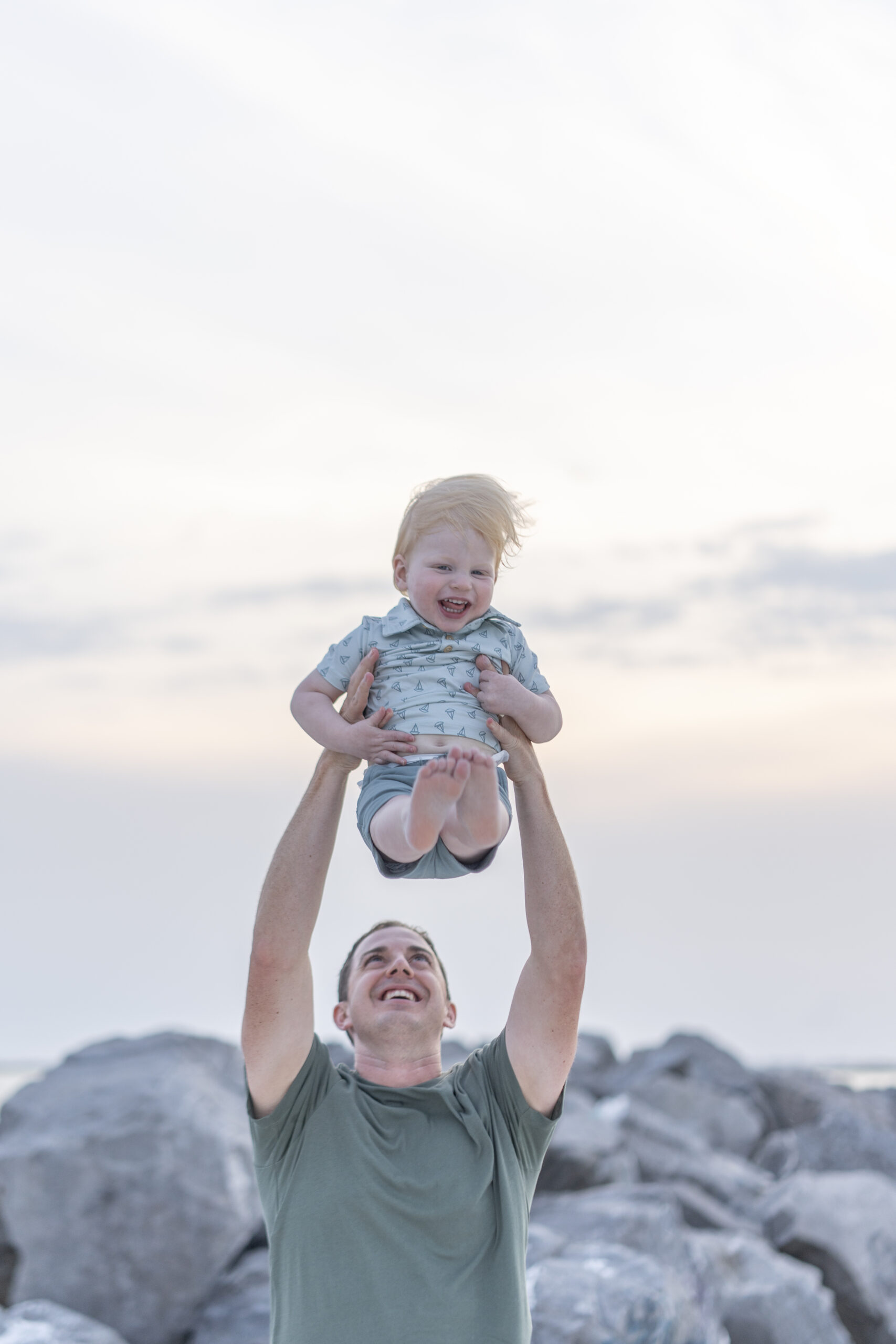 Dad lifting child into the air with joy at the Destin Jetties with clear blue water behind.
